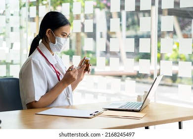 An Asian Female Doctor Works At A Hospital Desk, Recommending Convenient Online Services To Patients And Holding A Pill Bottle With Health Care Advice. Disease Prevention Concept