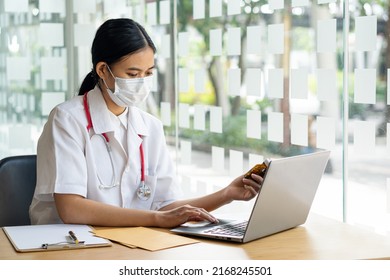 An Asian Female Doctor Works At A Hospital Desk, Recommending Convenient Online Services To Patients And Holding A Pill Bottle With Health Care Advice. Disease Prevention Concept