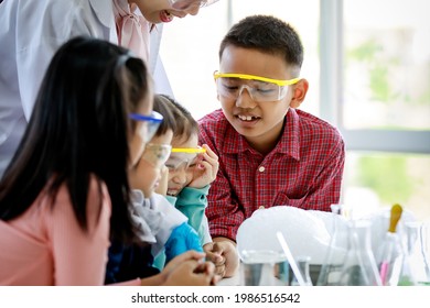 Asian Female Elementary School Science Teacher In Lab Coat Holding Colorful Laboratory Equipment In Hands Teaching Little Boy And Girls Wear Safety Goggles How To Use Them In Chemistry Experiment.