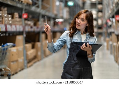 Asian female employee walks with a tablet to count inventory and look to buy raw materials in stock on a shelf in a warehouse. - Shutterstock ID 2158850567