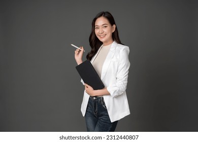 Asian female executive with long hair Smiling and giving outstanding eyes Working with a tablet and a pen wearing a white suit and stand to take pictures with a gray scene in the studio - Shutterstock ID 2312440807