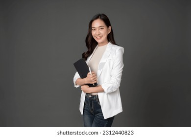 Asian female executive with long hair standing hugging a tablet for work, holding a pen, with a beautiful smile. wearing a white suit and stand to take pictures with a gray scene in the studio - Shutterstock ID 2312440823