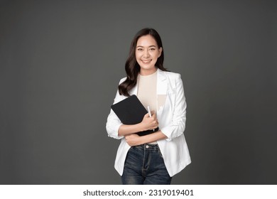 Asian female executive with long hair cute smile holding tablet and pen for work crossed arms, powerful posture Wearing a white suit, jeans and standing in a gray studio setting