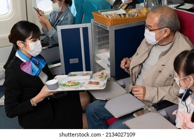 Asian Female Flight Attendant Serving An Airline Meal To Asian Senior Male Passenger And His Granddaughter, Placed On A Seat Tray Table. Everyone In The Plane Wearing A Face Mask To Protect Covid-19.