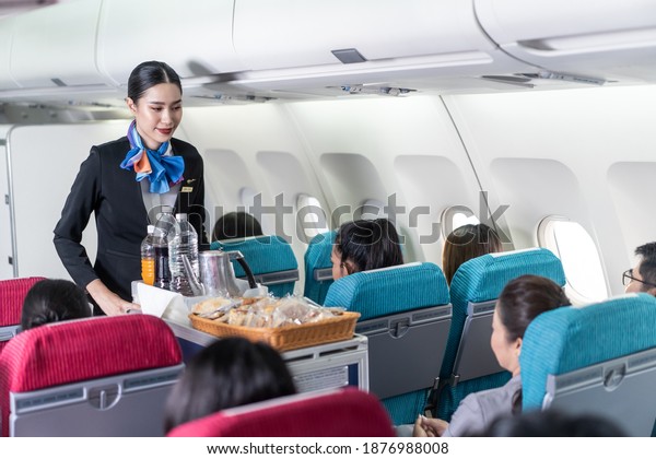 Asian female flight attendant serving food and\
drink to passengers on airplane. The cabin crew pushing the cart on\
aisle to serve the customer. Airline service job and occupation\
concept.