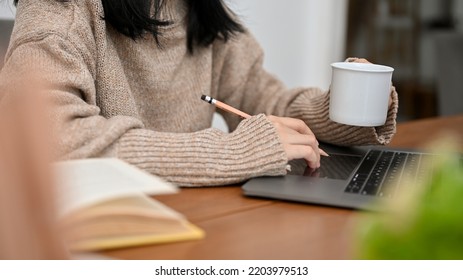 An Asian female freelancer or college student in comfy sweater working on her project, using laptop computer while sipping her morning coffee. cropped shot - Shutterstock ID 2203979513