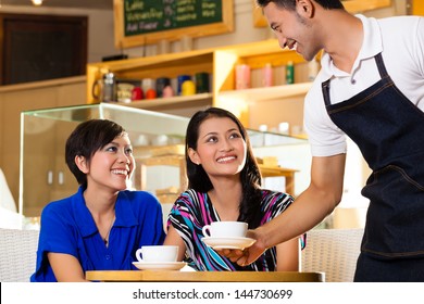 Asian female friends enjoying her leisure time in a cafe, drinking cappuccino and talking about some things, a Indonesian waiter serving the coffee
