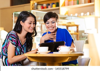 Asian Female Friends Enjoying Her Leisure Time In A Cafe, Drinking Coffee Or Cappuccino And Looking At Photos Or Emails On A Tablet Computer