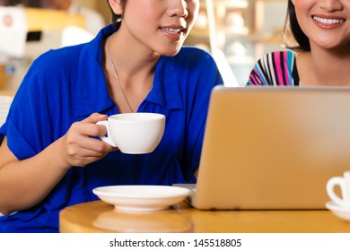 Asian Female Friends Enjoying Her Leisure Time In A Cafe, Drinking Coffee Or Cappuccino And Working On A Laptop Computer