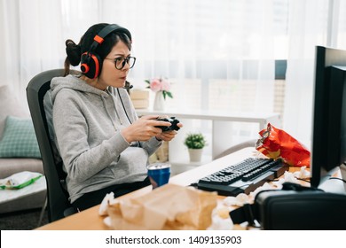 Asian Female Gamer Playing Computer Game With Controller In Headset Looking In Monitor With Relaxed Expression On Summer Break. Young Woman At Home Frowning With Junk Food Snack Trash