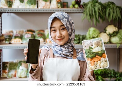 Asian Female Greengrocer Showing Vegetables And Showing A Phone Screen