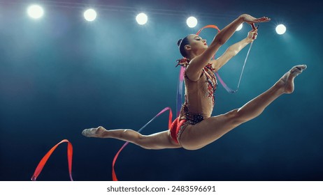 Asian Female Gymnast in Mid-air, Performing Dynamic Split Leap With Ribbon Under Bright Stadium Lights. Elegance, Flexibility, and Precision in an Exhilarating Rhythmic Gymnastics Routine. - Powered by Shutterstock