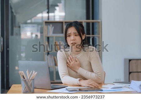An Asian female has a heart attack pain, and tension-sick employee who is feeling heartache while working on a laptop at the workplace at a desk in the office.