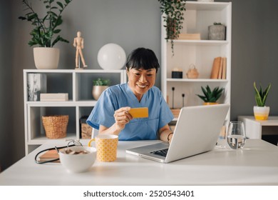 Asian female healthcare worker in blue scrubs holds a credit card during a session with her laptop in an organized office space, surrounded by bright indoor plants and neatly arranged shelves. - Powered by Shutterstock