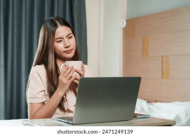 Asian female holding coffee cup while working on laptop in bedroom. Casual attire, long hair, thoughtful expression. Modern room design. Home office setup, natural light from window. - Powered by Shutterstock