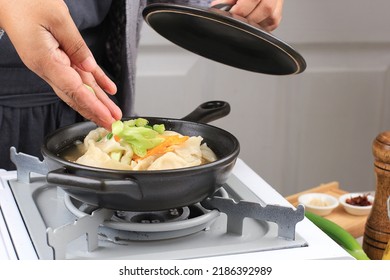 Asian Female Home Chef Cooking Dim Sum Soup Mandu Guk, Korean Dumpling Soup With Spring Onion Topping And Sliced Fried Egg Above The Stove. Put Sliced Spring Onion