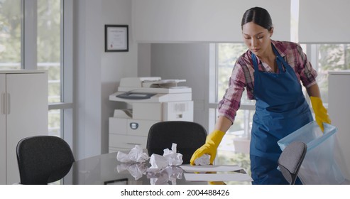 Asian Female Janitor Collecting Garbage In Bin Cleaning Office. Young Korean Maid In Apron And Gloves Cleaning Modern Empty Office Putting Trash In Bucket