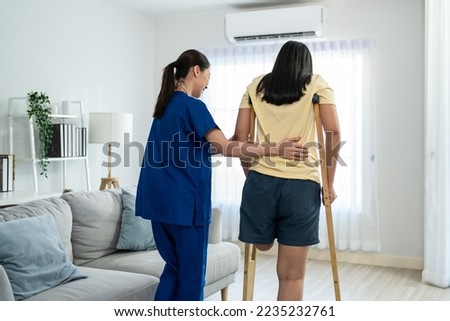 Asian female leg amputee doing physiotherapy with support from nurse. Attractive young girl legless patient practice walking slow with walker crutch with woman caregiver doctor in living room at home. Foto d'archivio © 