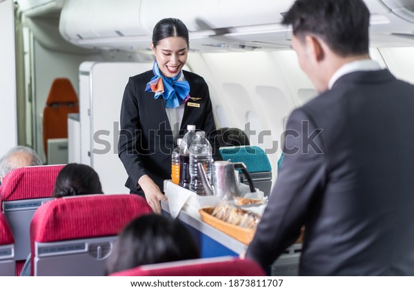 Asian female and male flight attendant serving\
food and drink to passengers on airplane. The cabin crew pushing\
the cart on aisle to serve the customer. Airline service job and\
occupation concept.