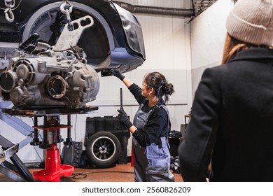 Asian female mechanic pointing at car engine part while installing gearbox in a car repair shop with customer - Powered by Shutterstock