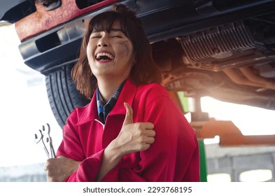 Asian female mechanic in red uniform enjoys working at auto workshop and gives thumbs up, working in auto service with lifted vehicle, car repair and maintenance - Powered by Shutterstock