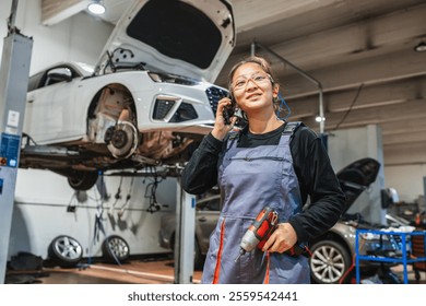 Asian female mechanic talking on phone while holding cordless drill with car on hydraulic lift in background in a garage - Powered by Shutterstock