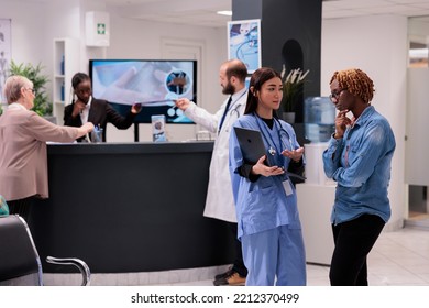 Asian Female Medical Staff Sharing On Laptop Screen Clinical Images With African American Patient At Hospital Reception. Young Receptionist Attending Doctor, Elderly Woman At Desk.