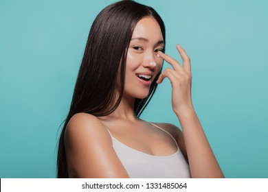 Asian female model applying moisturizer to her nose and looking at camera. Woman applying moisturizer cream on her face against blue background.