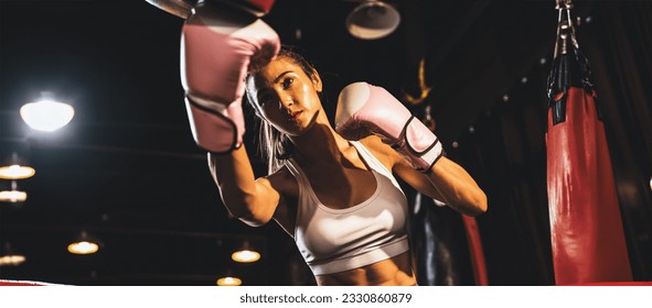 Asian female Muay Thai boxer punching in fierce boxing training session, delivering strike to her sparring trainer wearing punching mitts, showcasing Muay Thai boxing technique and skill. Spur - Powered by Shutterstock