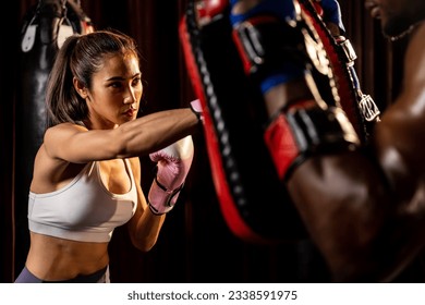 Asian female Muay Thai boxer punching in fierce boxing training session, delivering strike to her sparring trainer wearing punching mitts, showcasing Muay Thai boxing technique and skill. Impetus - Powered by Shutterstock