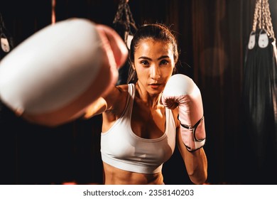 Asian female Muay Thai boxer punch fist in front of camera in ready to fight stance posing at gym with boxing equipment in background. Focused determination eyes and prepare for challenge. Impetus - Powered by Shutterstock