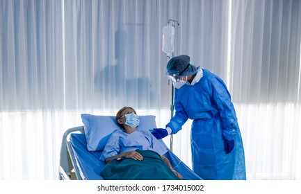 Asian Female Nurse In PPE Checks COVID-19 Infected Old Caucasian Patient In Quarantine Room. Patient Gets Infusion Therapy Care. Psychologist Gives Mental Advice To Old Virus Infected Patient At Bed