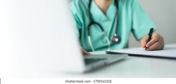 Asian Female Nurse Or Surgeon Doctor In Green Uniform Working On Laptop Computer And Writing Something Information Of Patient In Paperwork Checklist On Clipboard In Medical Room Of Hospital