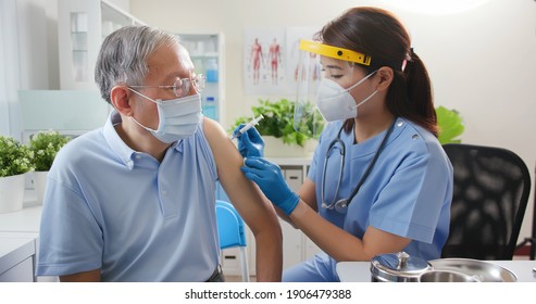 Asian Female Nurse Wearing Gloves And Isolation Mask Is Making A COVID-19 Vaccination In The Shoulder Of Senior Male Patient At Hospital