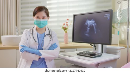 Asian Female Obstetrics And Gynecology Doctor Uses Computer Then Smiles At You - She Wearing Protective Face Mask To Prevent COVID19 With Uterus Utltrasonographic Picture On Screen In Hospital