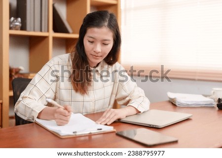 Asian female office worker, sitting signing documents related company work, was assigned project, has computer laptop is on desk because it's during working hours so we're ready work all time.