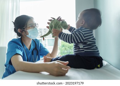 Asian Female Pediatrician Doctor Wearing Protective Face Mask Playing Dinosaur Doll With Her Little Baby Boy Patient Before Checkup In Medical Room At Hospital.