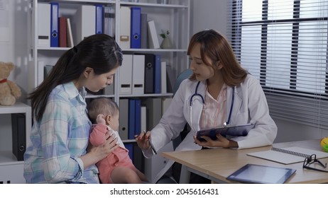 Asian Female Pediatrician In Her Office Is Observing The Lovely Baby Who’s In Her Mom’s Arms. Woman In White Coat Taking Note On Board During Well-baby Checkup. Real Moments