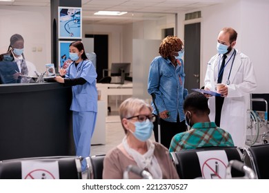 Asian Female Physician, African American Receptionist Checking Patient Itinerary At Hospital Reception Desk. Physiotherapist Doctor Accompanying Crutches Patient To Waiting Room, Explaining Health
