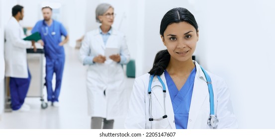 Asian Female Physician With Stethoscope Standing In Front View Of Hospital Lobby With Doctors In White Coats And Medical Staff. Working Day In Medical Clinic