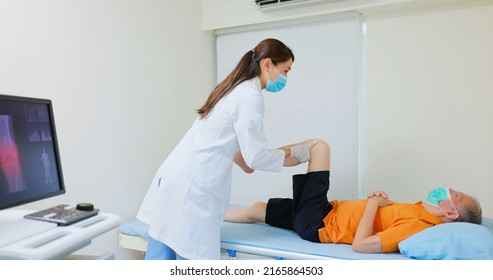 Asian Female Physiotherapist Is Helping Senior Elder Man Stretching His Hamstring And Doing Thigh Or Leg Rehabilitation In Exercise Room - He Lying On Massage Bed