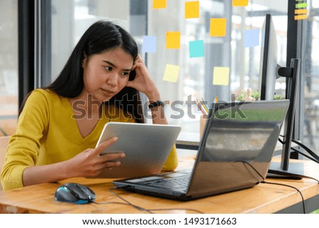 Asian female programmer, wearing a yellow shirt, looking at laptop screen, holding a tablet and pencil. She appeared happy.