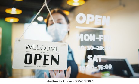 Asian Female Restaurant Owner Wear Face Mask, Flip The Shop's Sign Placard From Closed To Open. Business Reopen After Covid-19 Coronavirus Pandemic, Covid Relief, Or New Normal Lifestyle Concept