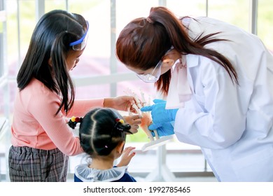 Asian Female Scientist Teacher Wears Safety Goggles White Lab Coat Rubber Gloves Teaching Little Curious Preschool Girls Observing Studying On Human Anatomy From Hand Skeleton Model In Laboratory.