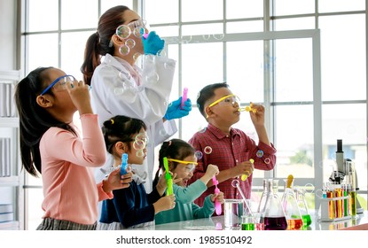 Asian female scientist teacher in white lab coat having fun playing blowing soap bubbles with little innocent happy pre-elementary boy and girl children wears safety goggles in school laboratory.