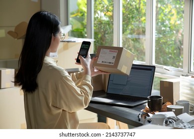 Asian Female Small Business Owner Using Mobile App On Smartphone Checking Parcel Box. Warehouse Worker, Seller Holding Phone Scanning Retail Drop Shipping Package Postal Parcel Bar Code.