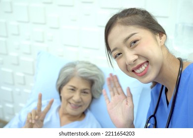 Asian Female Specialist Doctor Using A Smartphone For Video Call To Let Asian Senior Elder Patient Talking To Family While Recovering In The Hospital. Nurse Using Selfie Video Call On A Smartphone.