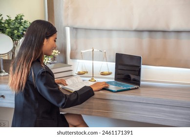 Asian female student, law office intern, is a lawyer, sitting at a desk with a scale, reading a book, researching legal issues, typing on a laptop. - Powered by Shutterstock