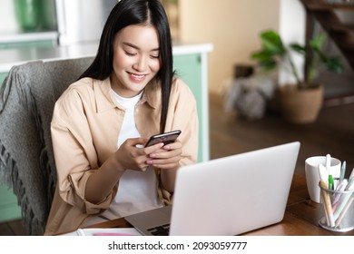 Asian Female Student Looking At Mobile Phone, Sitting At Home With Laptop, Register Online Course On Laptop, Working Remotely