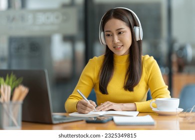 Asian female student or office worker studying online using headphones and laptop taking notes in notebook sitting at coffee shop table relaxing and changing places to work relax.